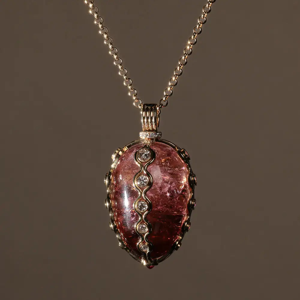 A pendant featuring a large, oval-shaped pink tourmaline stone set in an intricate gold-toned metal frame with a delicate zigzag pattern of embedded diamonds along its center.