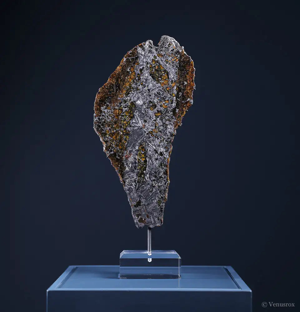 A rugged, irregularly shaped meteorite with a dark metallic surface speckled with patches of orange and green oxidation rests on an acrylic display stand.
