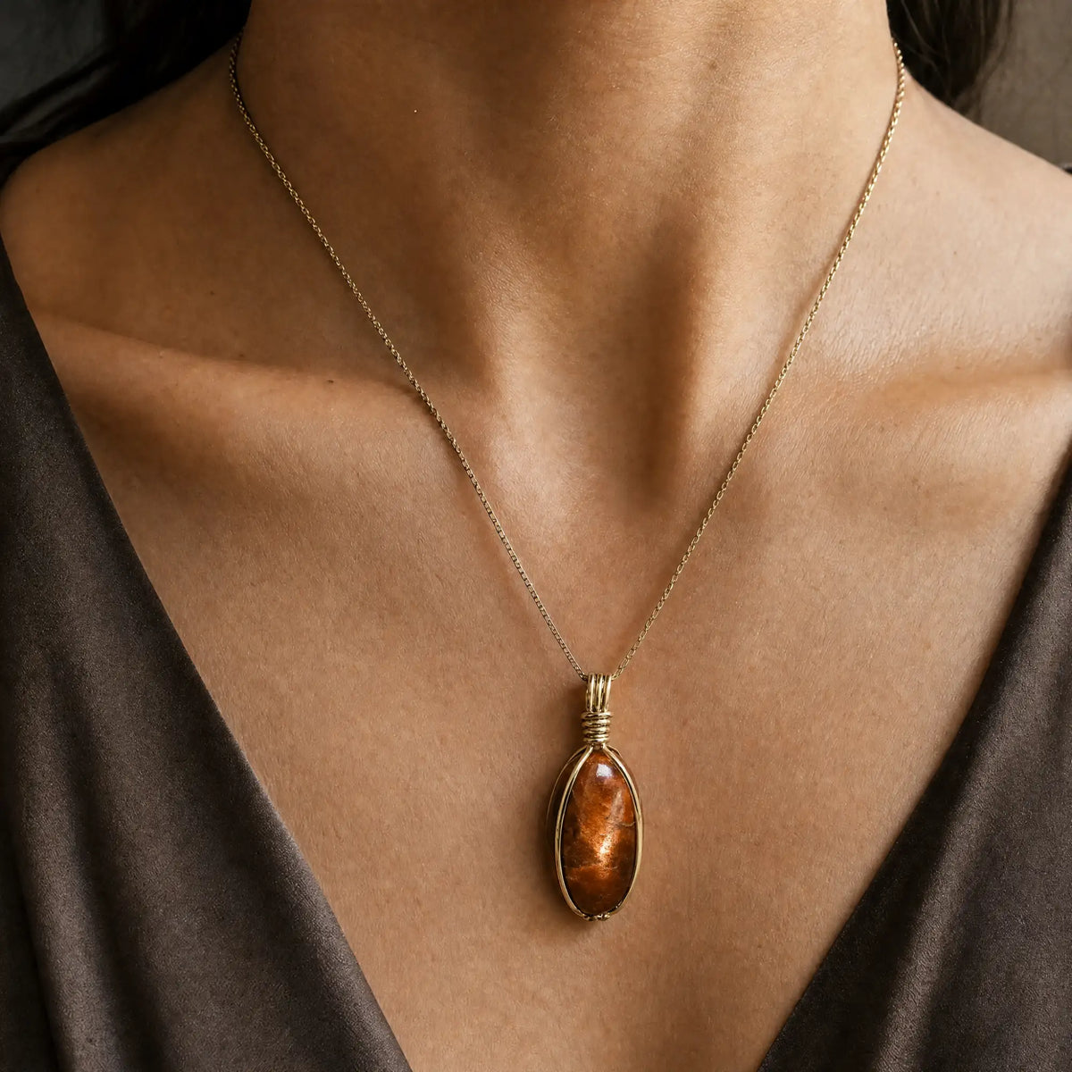 A warm amber-colored, oval-shaped pendant with a wire-wrapped gold-toned metal setting hangs from a delicate gold chain.